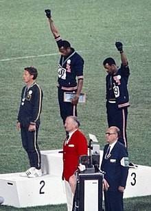 Tommie Smith et John Carlos, n’ayant pu récupérer qu’une paire de gants avant la cérémonie, se la partagent. Sur la deuxième marche du podium, l’Australien Peter Norman arbore lui aussi le badge de l’Olympic Projet for Human Rights, en signe de solidarité. Source : Angelo Cozzi, domaine public, via Wikipedia.