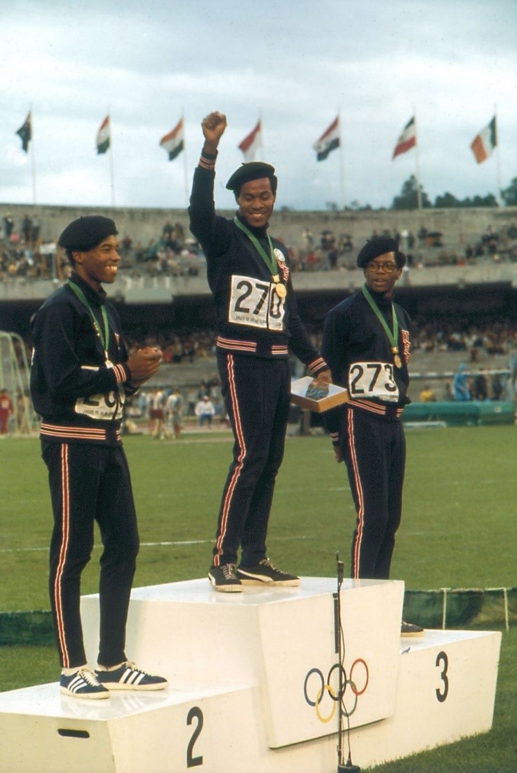 Lee Evans (au centre), Larry James (à gauche) et Ron Freeman (à droite) portent un béret, symbole associé aux Black Panthers. Lee Evans lève le poing, comme Tommie Smith et John Carlos la veille. Leur sourire contraste en revanche avec l’attitude solennelle qui prévalait chez les deux bannis. Source : compte X de l’USA Track and Field
